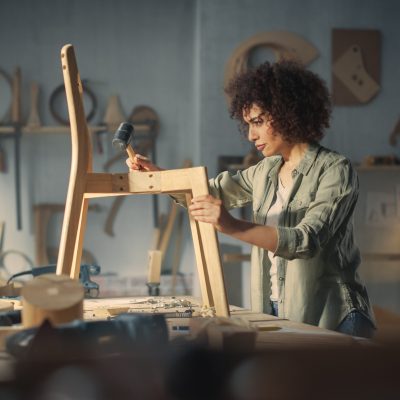 Young Woodworker Checking the Layout Manual of a Stylish Handmade Wooden Chair. Talented Female Furniture Designer Working in a Workshop in a Creative Loft Space with Tools and Equipment.